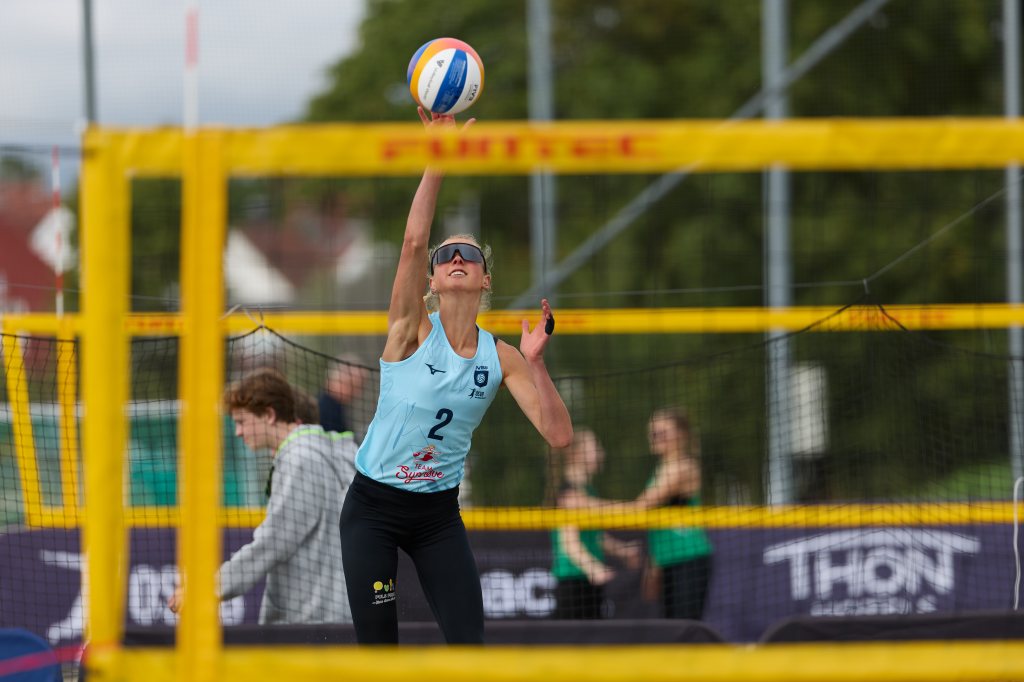 En kvinne i sandvolleyball står klar til å serve, iført solbriller og sportsdrakt med nummer 2. I bakgrunnen er det andre spillere og trenere, med volleyballnett og gule sperringer rundt banen.
