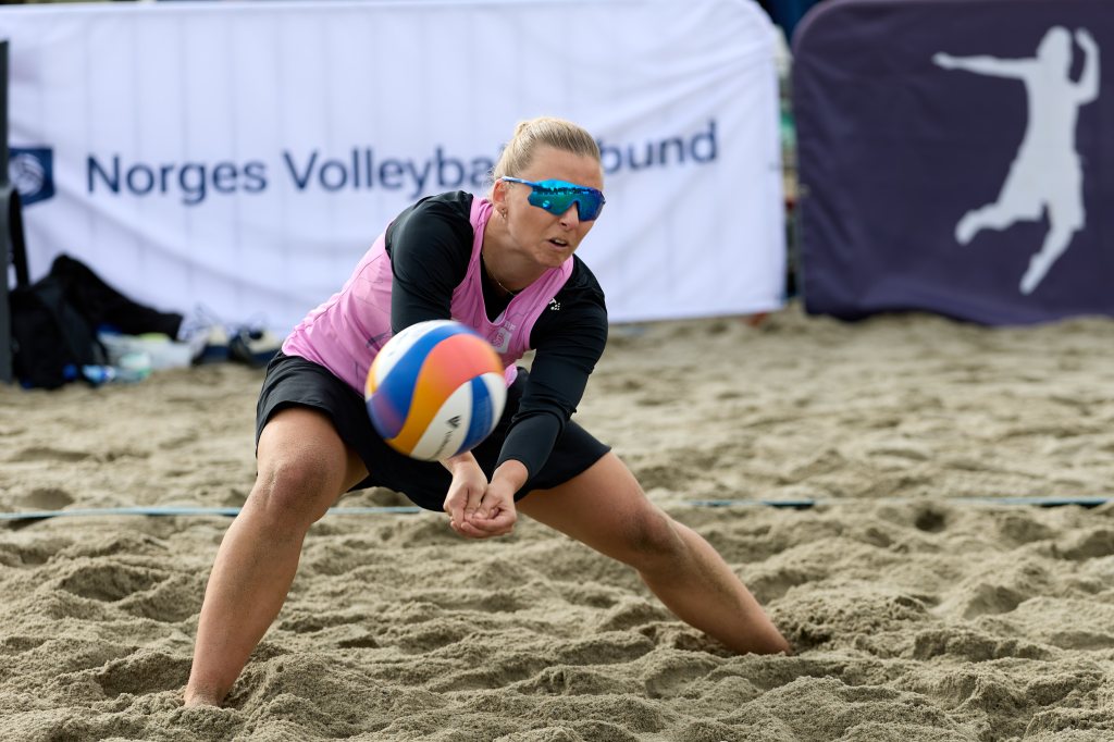 En strandvolleyballspiller i aksjon, iført solbriller og sportsklær, klarer å ta imot ballen på sandbanen.