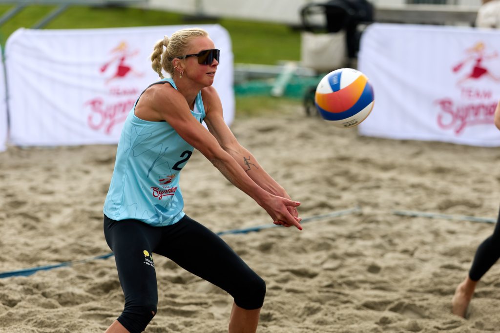 En kvinne i sandvolleyball, iført en lys blå topp med nummer 2, spiller ball. Hun har solbriller på og viser fokusert uttrykk mens hun setter seg for å ta imot ballen.