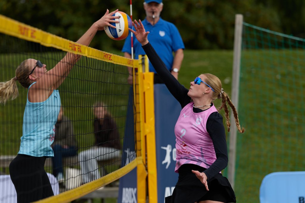 To kvinner spiller volleyball over nettet, en i blå topp og den andre i rosa. Begge bruker solbriller.