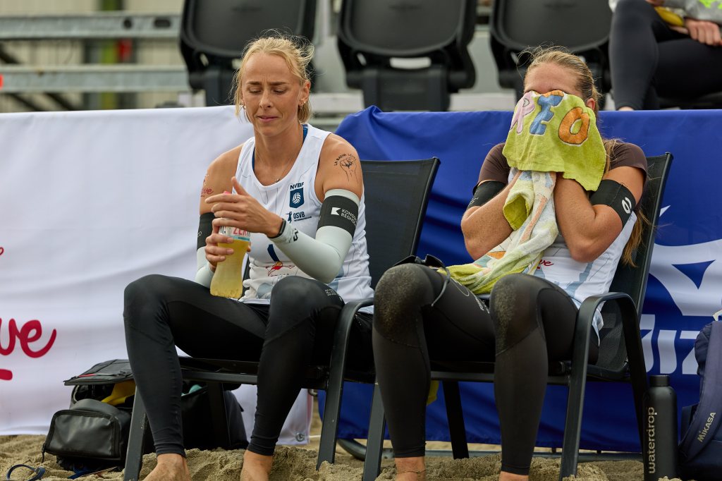 To kvinnelige volleyballspillere sitter på stoler i sanden, den ene drikker fra en flaske mens den andre dekker ansiktet med et håndkle.