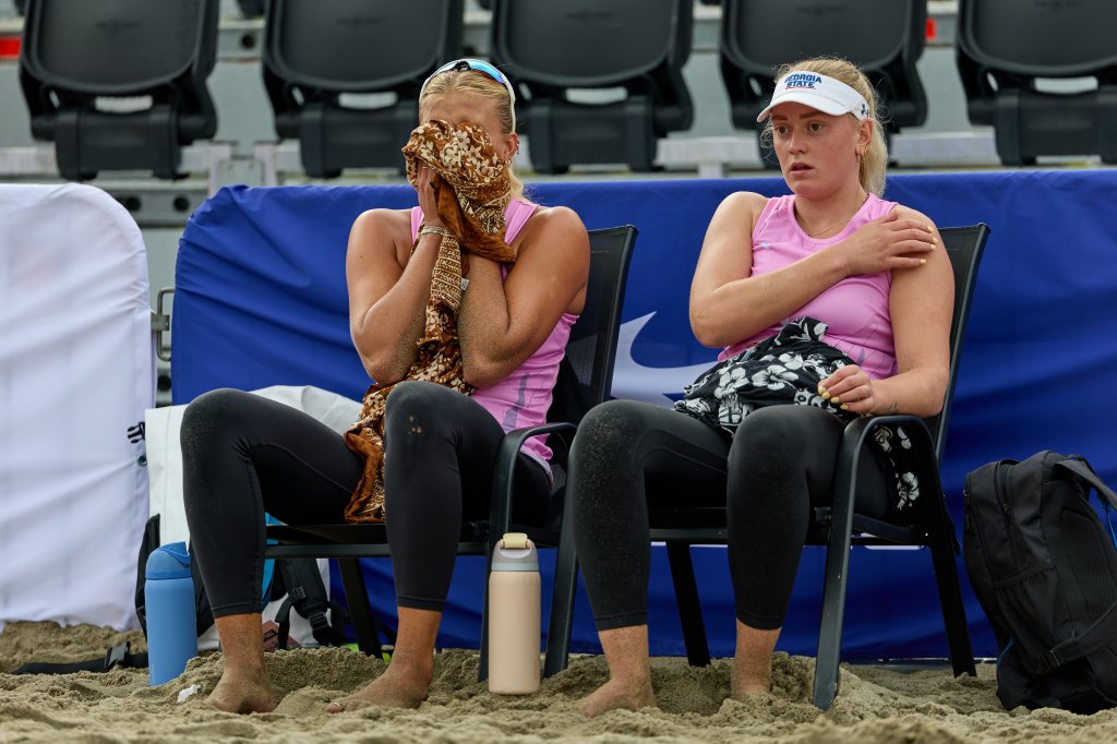 To kvinner sitter på strandstoler iført rosa treningsklær. Den ene kvinnen dekker ansiktet med en scarf, mens den andre ser usikker ut. Begge har sand på føttene og er plassert på stranden med tribuner i bakgrunnen.