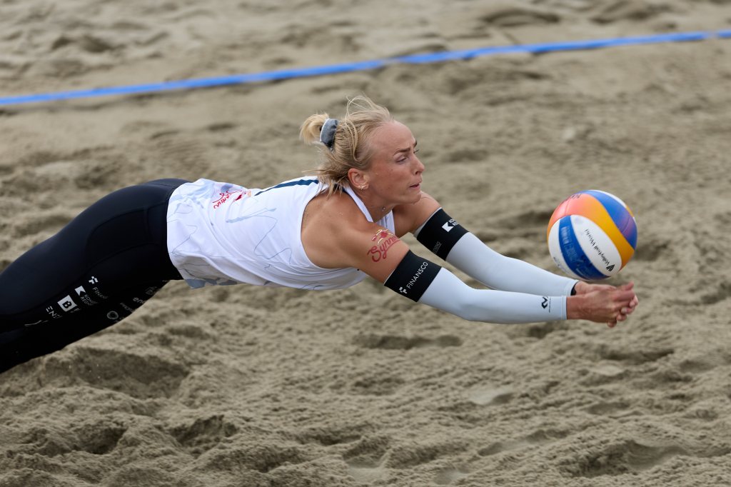 En sandvolleyballspiller i aksjon, som strekker seg etter ballen mens hun faller mot sanden.