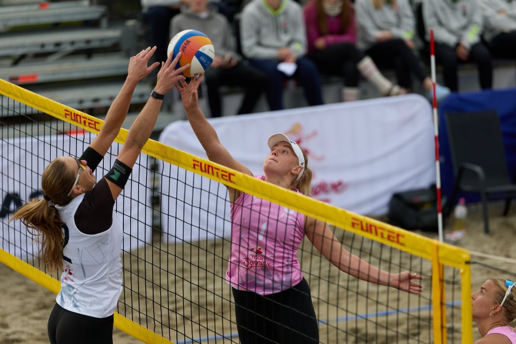 To kvinne spillere i en beachvolleyballkamp, der den ene prøver å blokkere ballen over nettet.