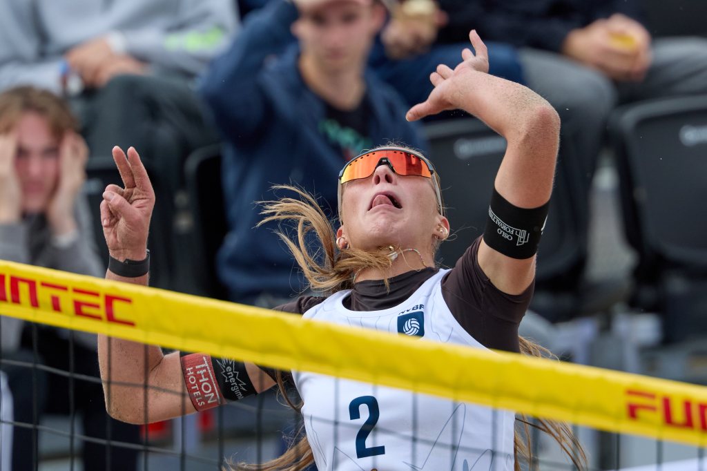 En kvinnelig volleyballspiller i aksjon, løfter armen for å slå ballen over nettet. Hun har på seg solbriller og har langt hår. I bakgrunnen er det tilskuere som følger med.