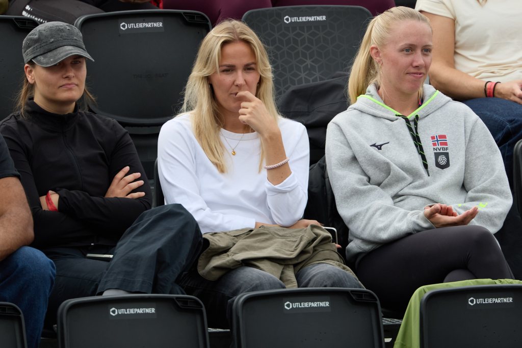 Tre kvinner sitter på tribunen, iført sporty klær, med fokusert uttrykk mens de ser på en sportsbegivenhet.