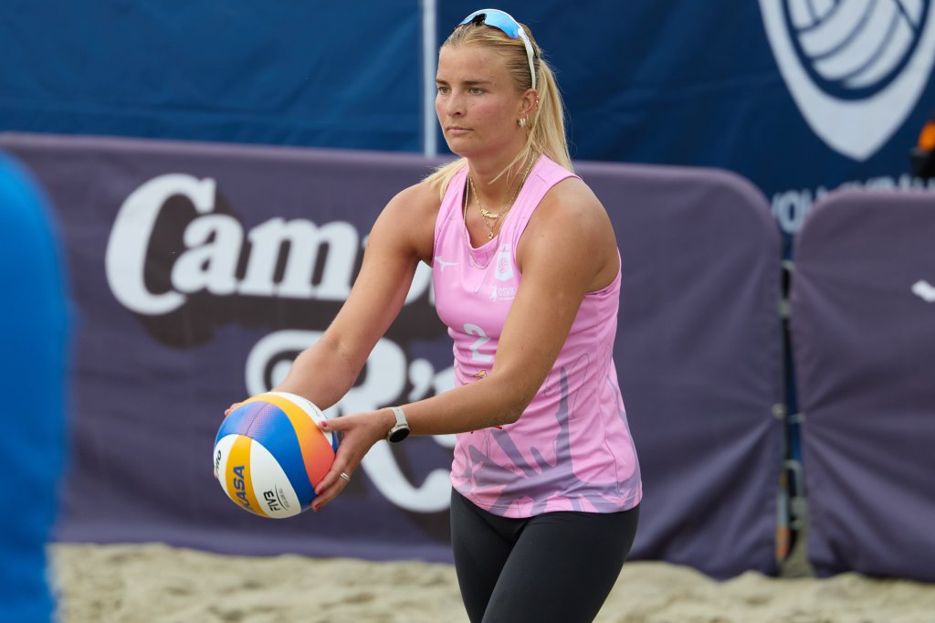 En kvinne med blondt hår iført en rosa sports-top og svarte treningbukser holder en volleyball på en sandvolleyballbane.