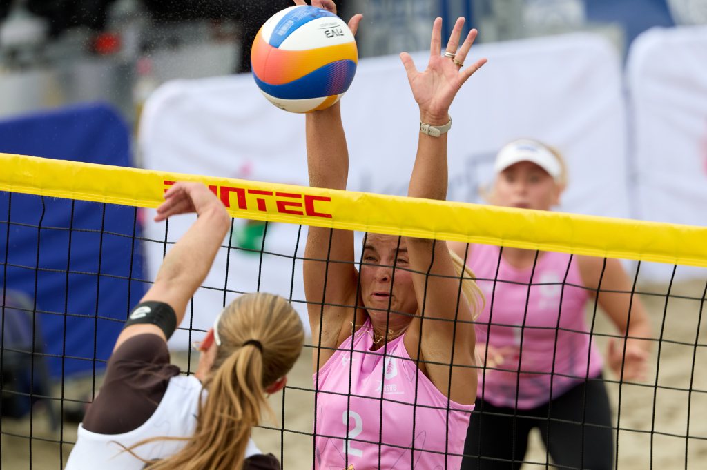 To kvinnelige beachvolleyballspillere i aksjon ved nettet. Den ene spilleren i rosa hever hånden for å blokkere et serveforsøk fra motstanderen.