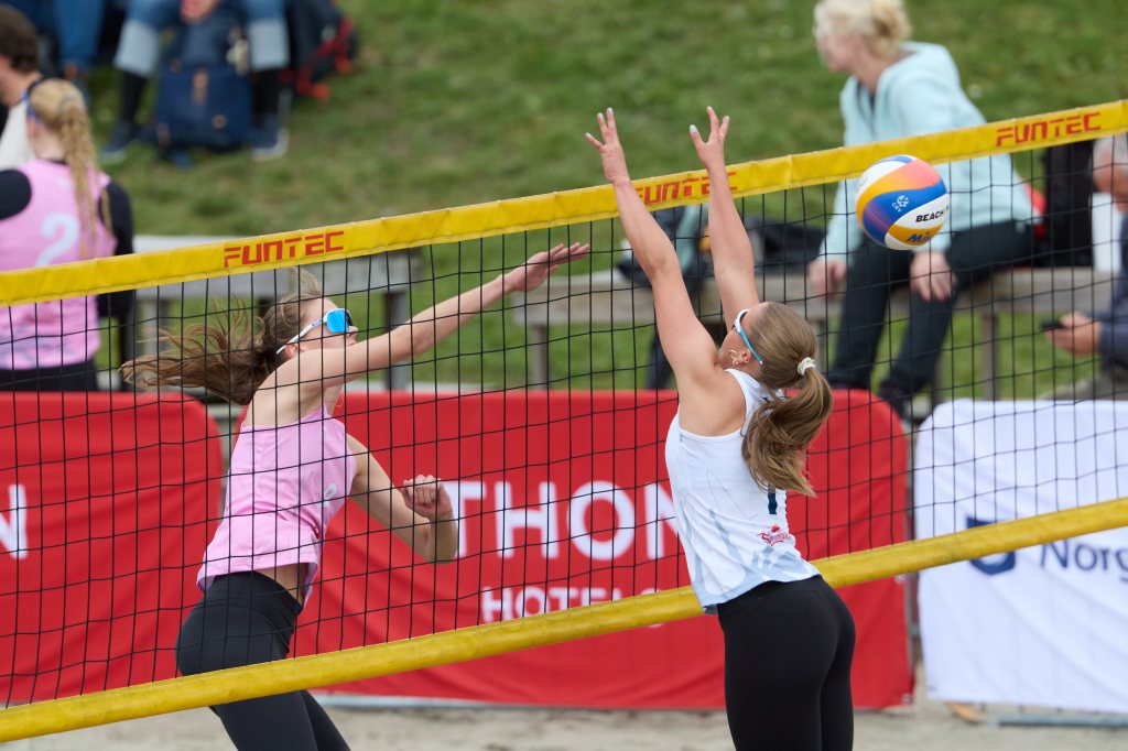 To kvinnelige volleyballspillere i aksjon på en sandbane, med en ball på vei over nettet. Den ene spilleren i rosa gjør et hopp for å blokkere, mens den andre i hvit forbereder et angrep.