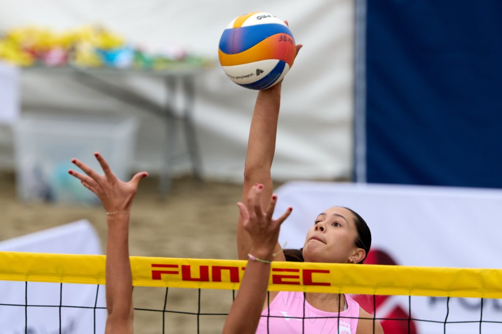 En sandvolleyballspiller hopper for å slå ballen over nettet, mens motstanderen prøver å blokkere.