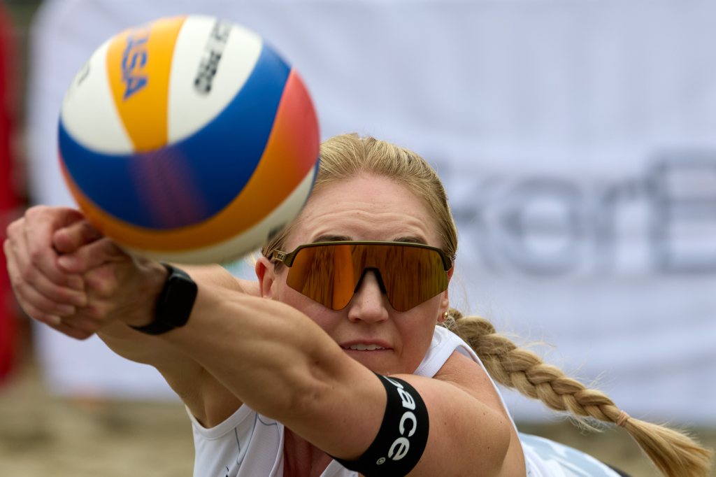 En kvinne spiller beachvolleyball og strekker seg etter ballen med solbriller på.
