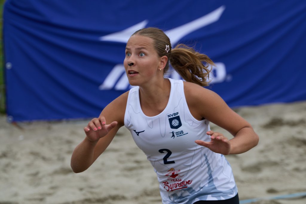 Ung kvinne som spiller volleyball på sandbane, iført hvit drakt med nummer 2. Hun ser opp mot ballen med fokusert uttrykk.