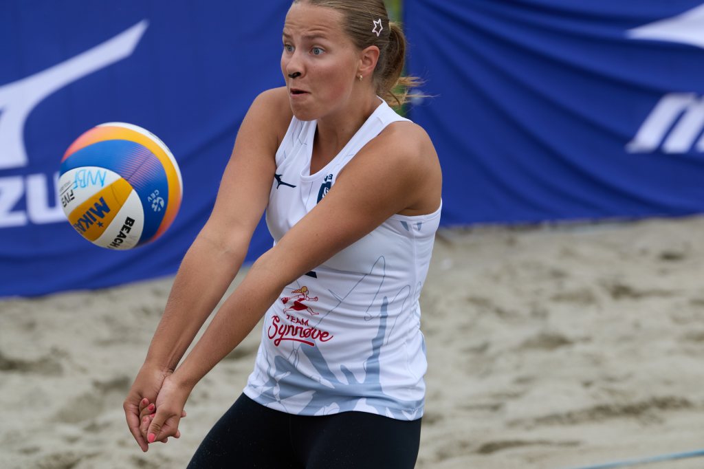 Dame spiller beachvolleyball, forbereder seg på å motta ballen, med en intens uttrykk. Sand og blå bakgrunn fra sponsorbanner er synlig.