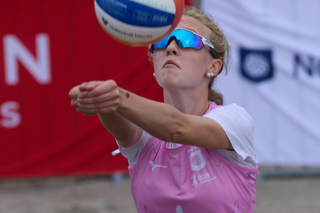 En kvinne spiller sandvolleyball og ser opp mot ballen mens hun forbereder seg på å motta den. Hun har på seg rosa sportstøy og solbriller.