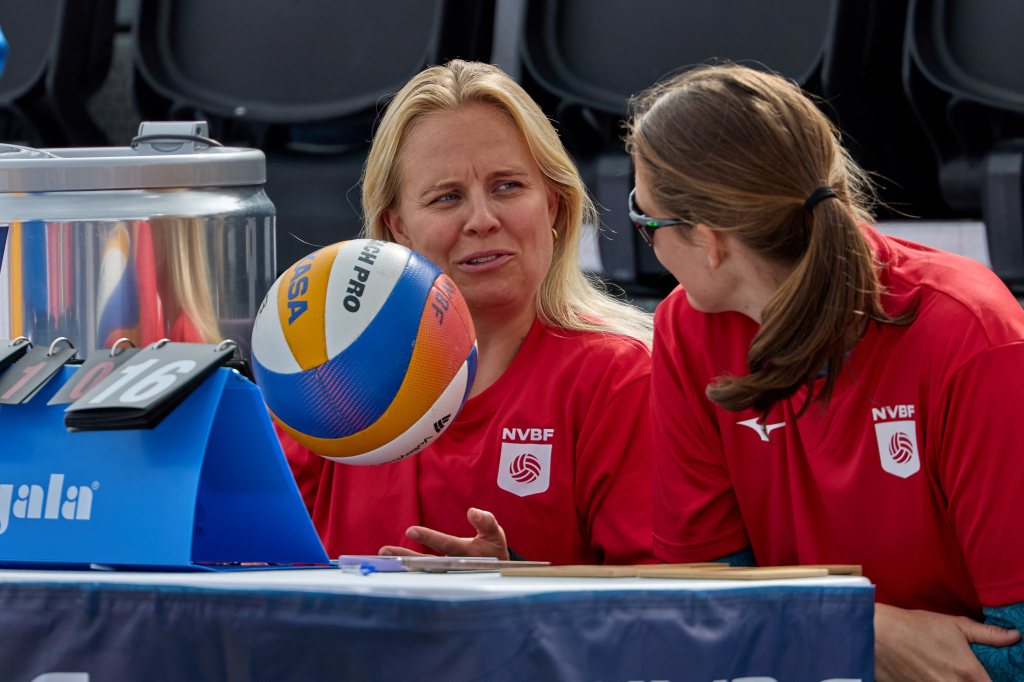 To kvinner i røde t-skjorter sitter ved et bord med en volleyball og diskuterer. I bakgrunnen er det en klokke og stoler.
