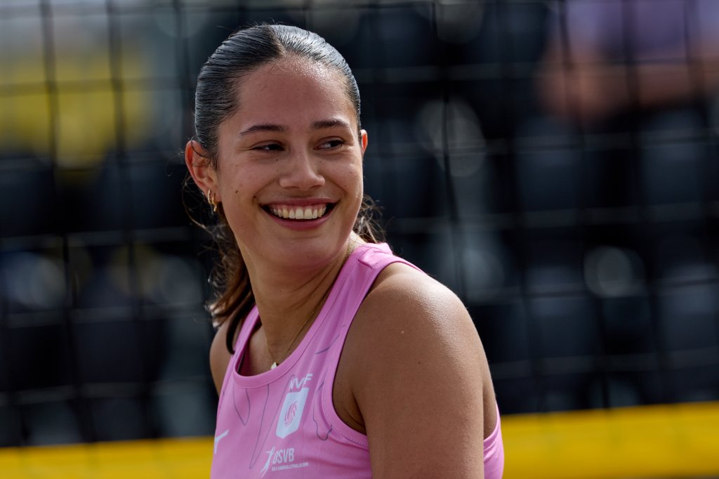 En smilende kvinne iført rosa sportsdrakt, står foran et volleyballnett.