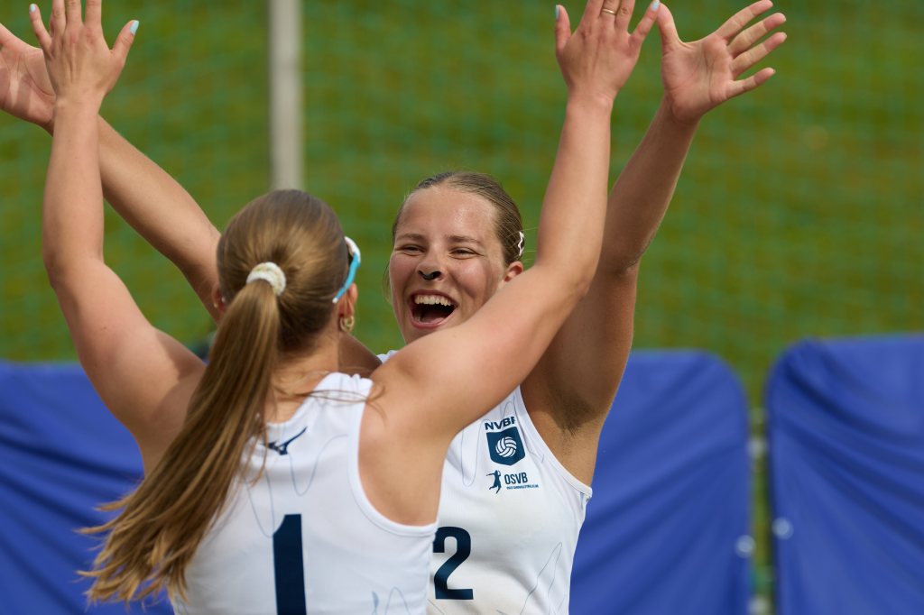 To kvinner jubler sammen etter å ha vunnet et poeng i sandvolleyball, iført hvite drakter.