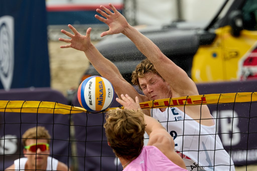 To beachvolleyballspillere i aksjon under en kamp, hvor en spiller prøver å blokkere ballen over nettet.