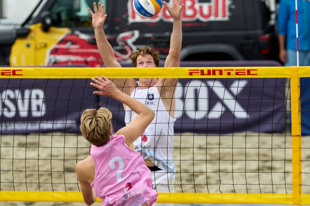 To beachvolleyballspillere i aksjon på en sandbane, en spiller hopper for å blokkere ballen mens den andre spilleren forbereder seg til å slå den.
