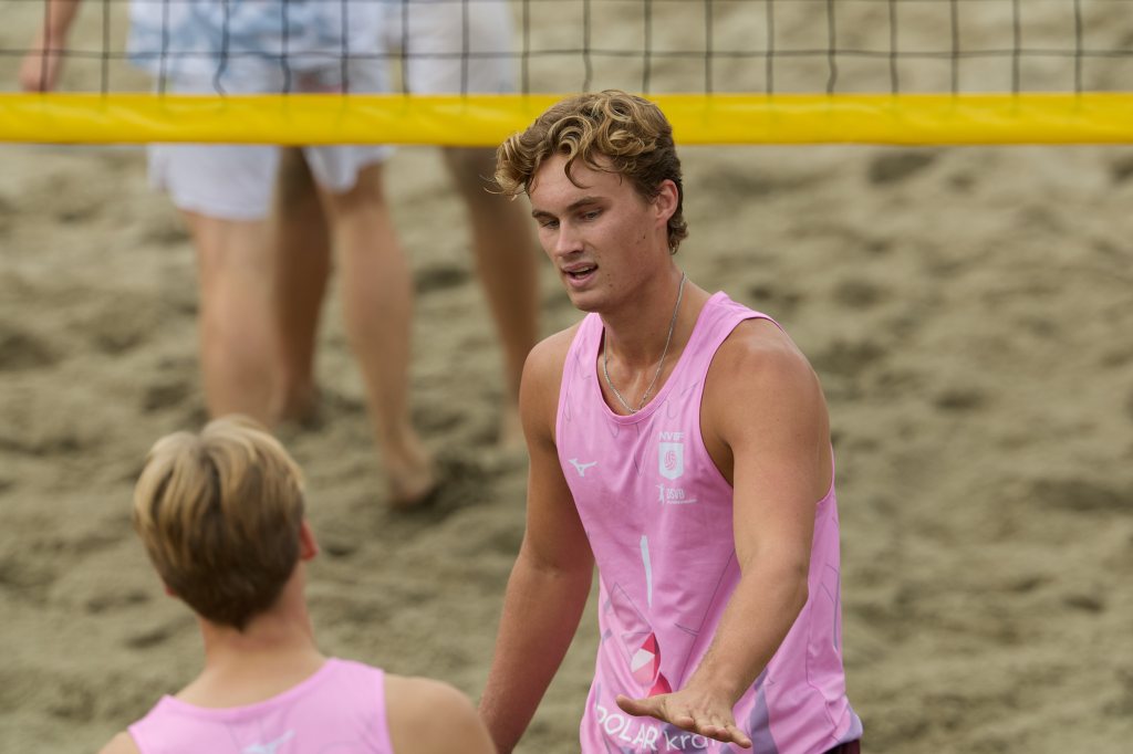 To unge menn i rosa treningstopper snakker sammen på en sandvolleyballbane, med et volleyballnett bak dem.