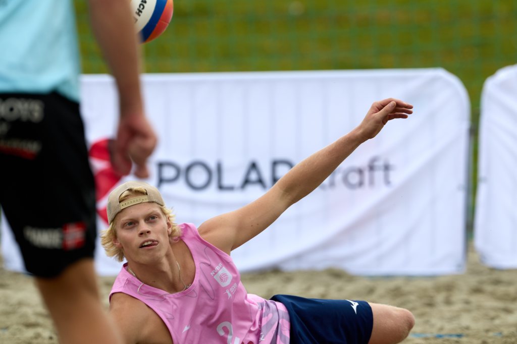 En ung mann i rosa sportsdrakt prøver å redde en volleyball under en strandvolleyballkamp.