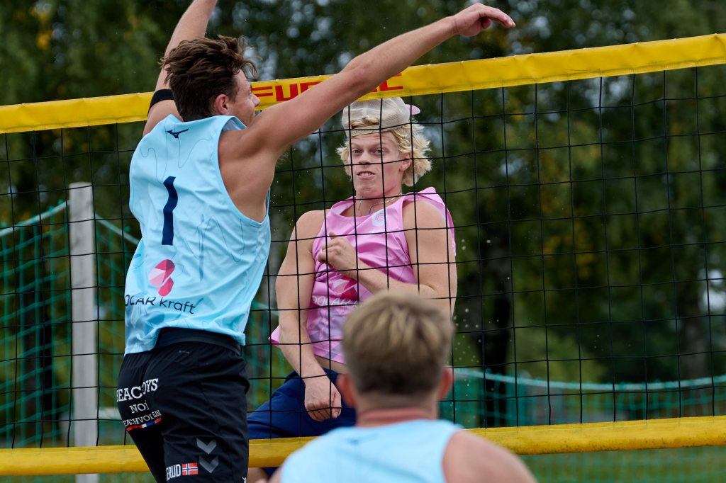 To volleyballspillere i aksjon, med en spiller i blå drakt som hopper for å blokkere et angrep fra en spiller i rosa drakt. Nettet mellom dem og en annen spiller delvis synlig i bakgrunnen.