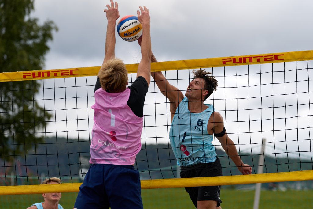 To volleyballspillere i aksjon, en spiller i rosa drakt hopper for å blokkere en ball som blir slått av en spiller i lyseblå drakt, med nett mellom dem.