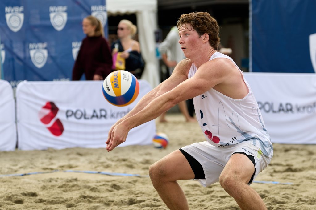 En ung mann i trening med beach volleyball, som forbereder seg på å ta imot ballen på en sandbane. I bakgrunnen er tilskuere og bannere synlige.