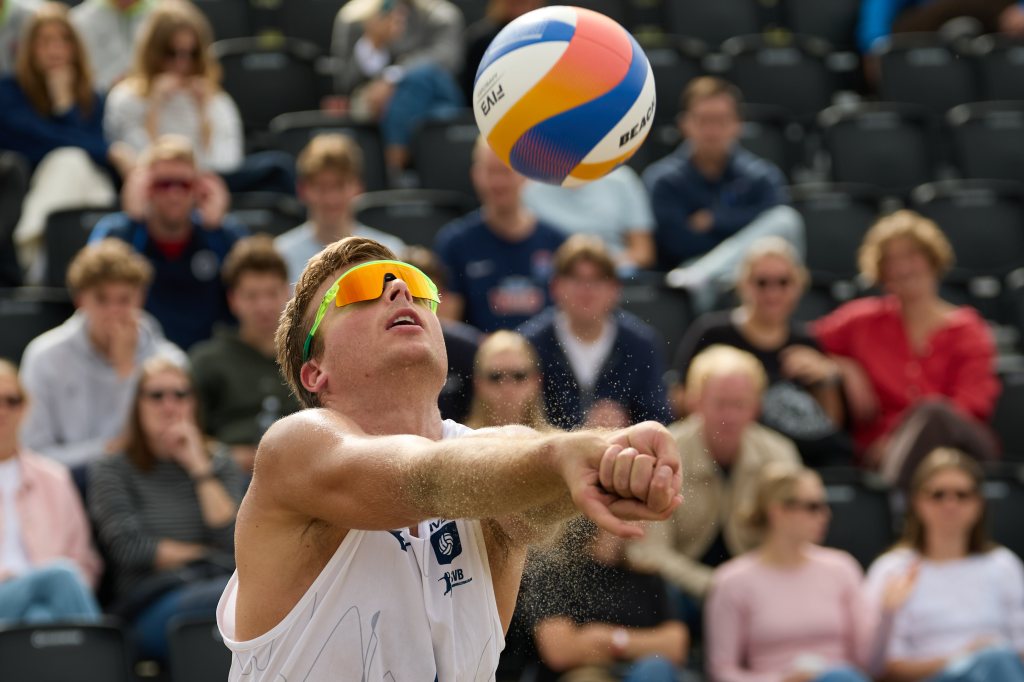 En volleyballspiller i solbriller mottar en volleyball i sand med publikum i bakgrunnen.