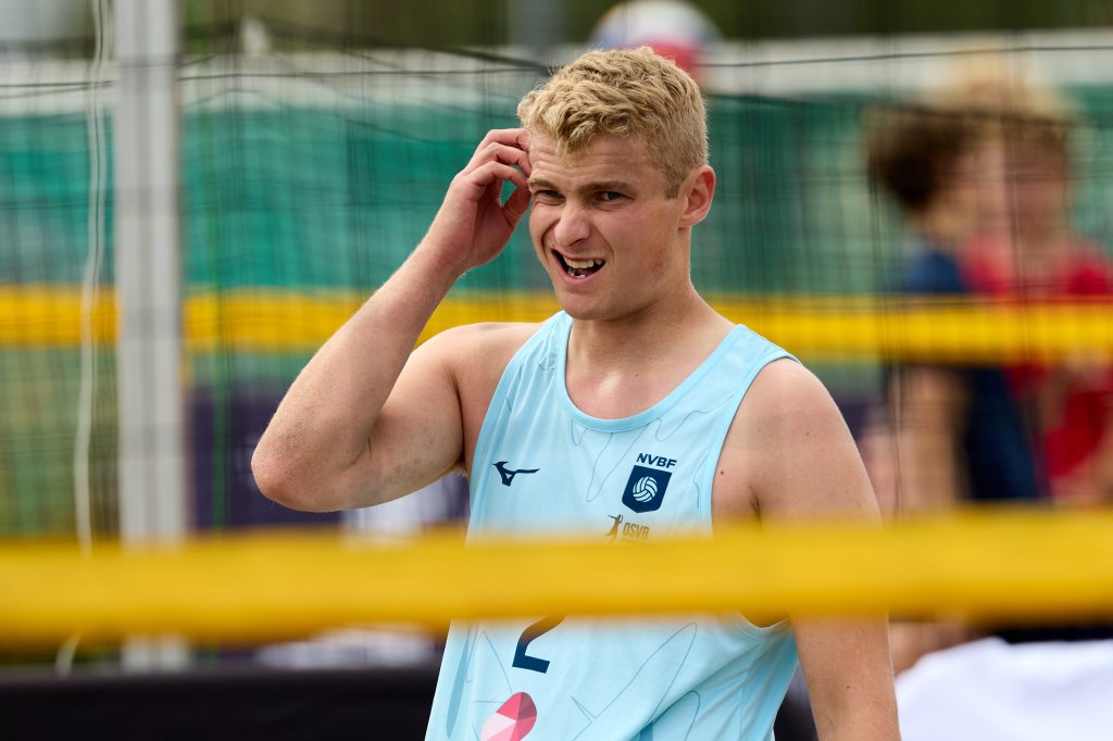 En ung mann i en lys blå treningstrøye står på en sandvolleyballbane og smiler mens han rører ved håret sitt.