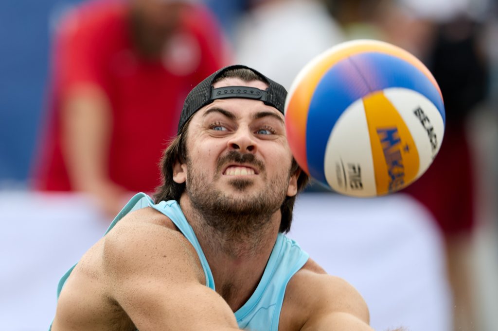Mann iført blå t-skjorte og caps, som kjemper for å ta imot en volleyball i et beachvolleyballspill.
