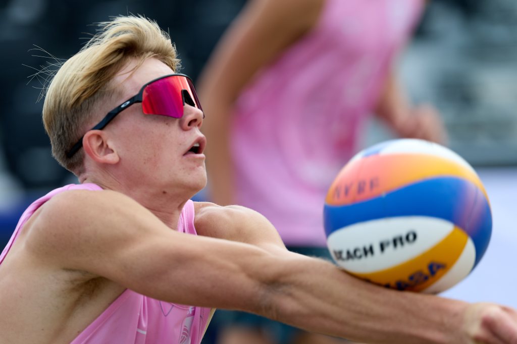 En ung mann i rosa sportsdrakt og solbriller spiller volleyball, fokusert på ballen som kommer mot ham.