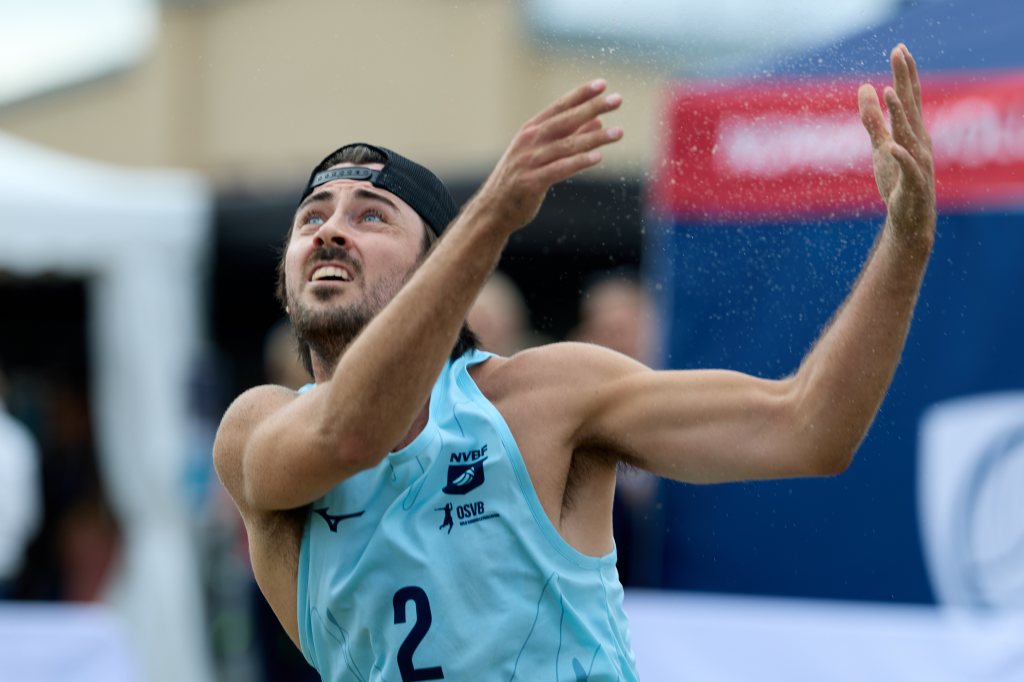 En mann i blå sportsdrakt som spiller volleyball på sand, ser opp mot ballen med fokus.