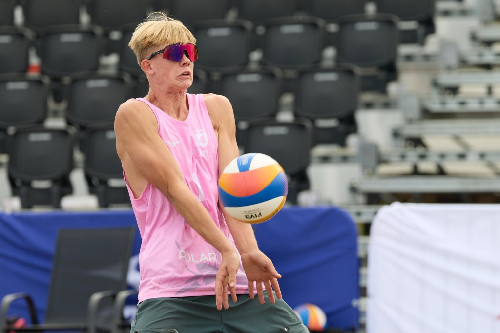 En volleyballspiller i rosa singlet fanger en ball med en intensitet på en sandbane, omgitt av tomme tribuner.