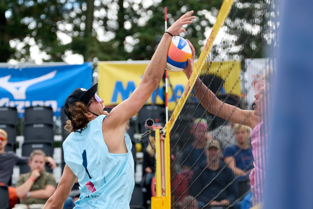 En spiller i lys blå drakt hopper for å blokkere et volleyballslag på en strandvolleyballbane.