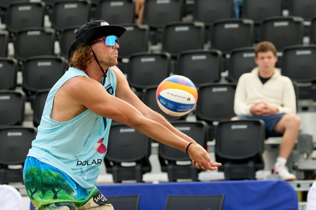 En mann i blått treningsantrekk og svart hatt spiller volleyball på en utendørs arena, med en tilskuer i bakgrunnen.