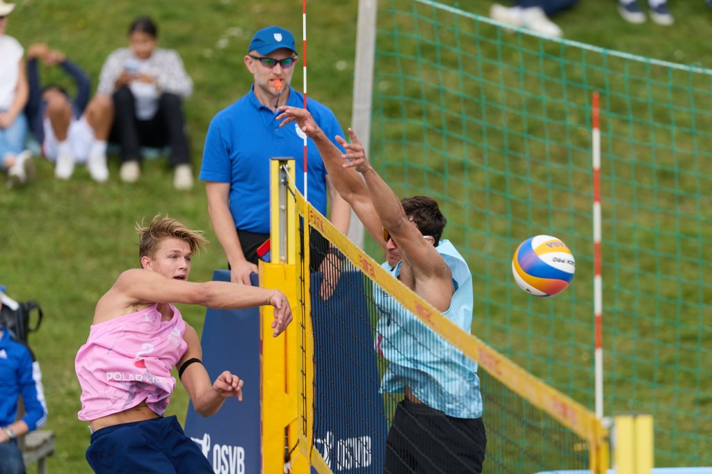 To unge menn spiller beachvolleyball, én i rosa skjorte som slår ballen, og den andre i blå skjorte som prøver å blokkere.