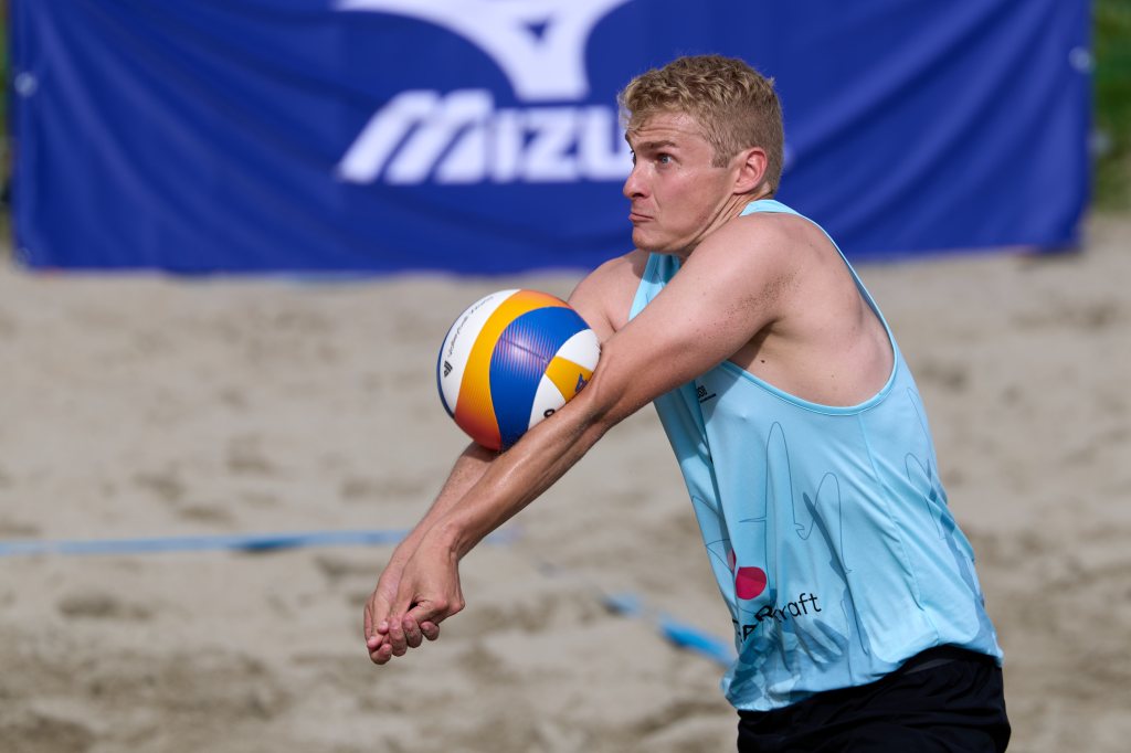 En mann spiller beachvolleyball, iført en lys blå singlet, mens han forbereder seg på å ta imot ballen på en sandbane.