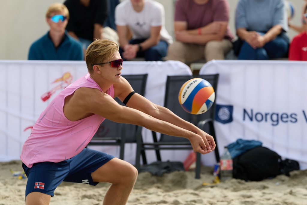En ung mann i rosa treningstrøye og shorts spiller volleyball på sand. Han utfører en mottak av ballen med fokus. I bakgrunnen er tilskuere som ser på kampen.