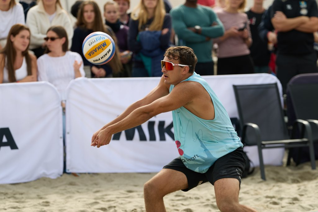 En volleyballspiller i en blå tanktop forbereder seg på å ta imot ballen under en strandvolleyballkamp, med publikum i bakgrunnen.