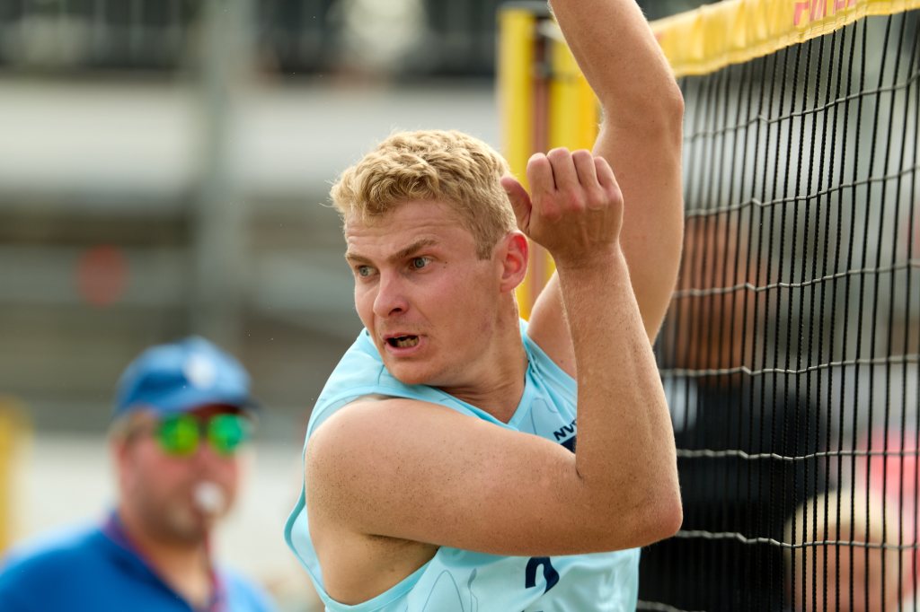 En ung mann som spiller beachvolleyball, forbereder seg på å slå ballen over nettet. Han ser fokusert og intens ut, iført en lys blå treningsoverdel.