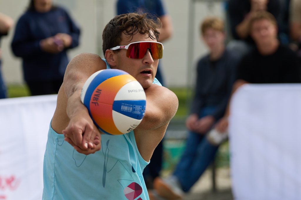 En ung mann i solbriller og lys blå tanktopp som spiller volleyball på en utendørsbane, mens tilskuere følger med i bakgrunnen.