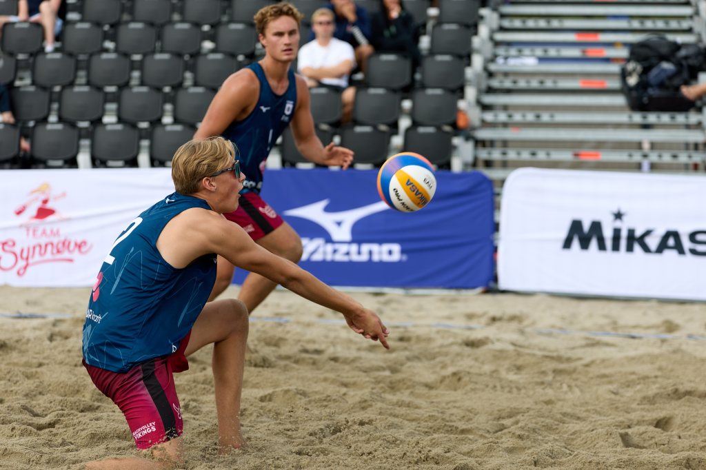 To beachvolleyballspillere i aksjon på sandbanen, med en ball i luften. Den ene spilleren er i ferd med å gjøre et mottak.