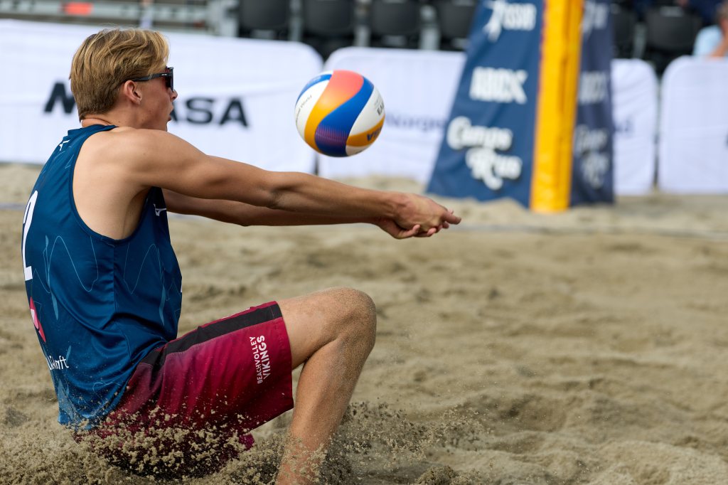 En sandvolleyballspiller sitter på bakken og forbereder seg på å slå et ball som kommer mot ham.