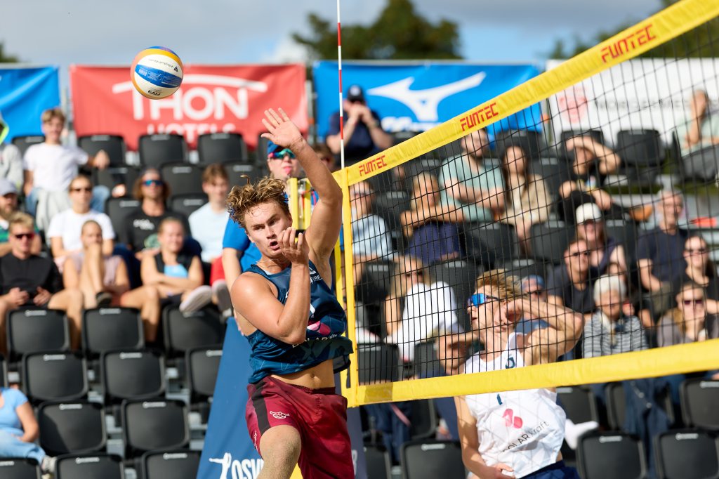En mann som spiller strandvolleyball, hopper for å gjøre en smasher, med publikum som ser på i bakgrunnen.