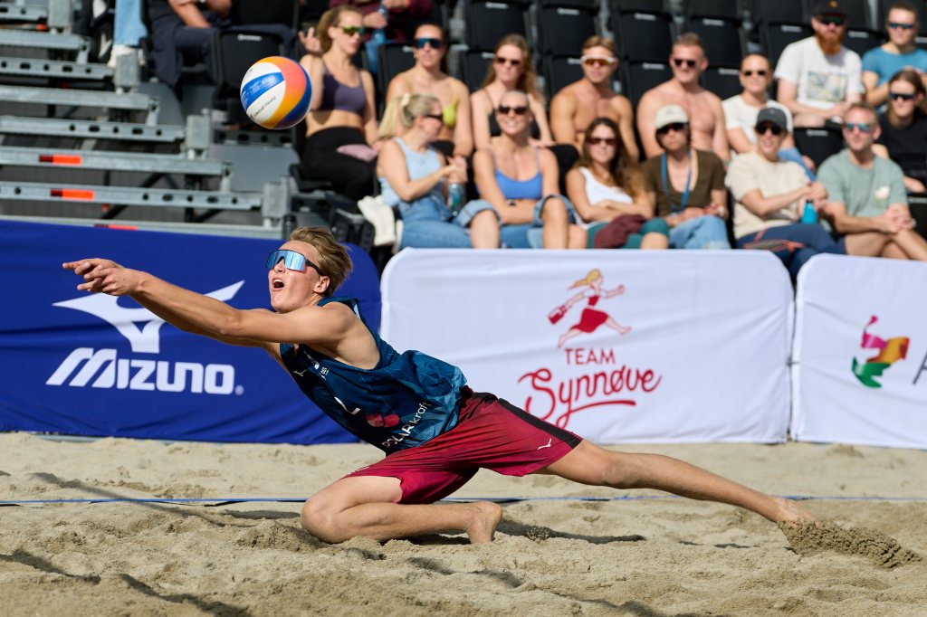 En ung mann i sportsbekledning utfører et dyktig stup for å fange en volleyball på sandstranden, med tilskuere i bakgrunnen.