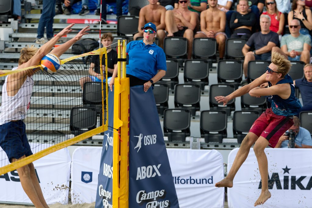 To unge menn spiller sandvolleyball i en konkurranse, med en dommer i bakgrunnen. Den ene spilleren hopper for å slå ballen over nettet.