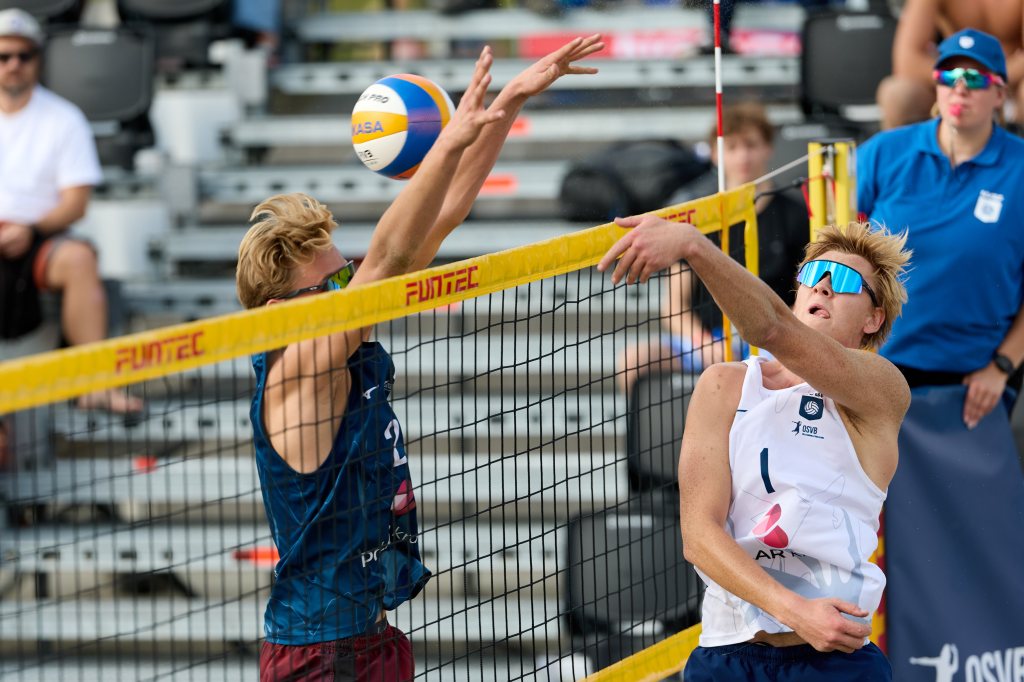 To strandspillere i sandvolleyball i aksjon, der en spiller hopper for å blokkere ballen over nettet.