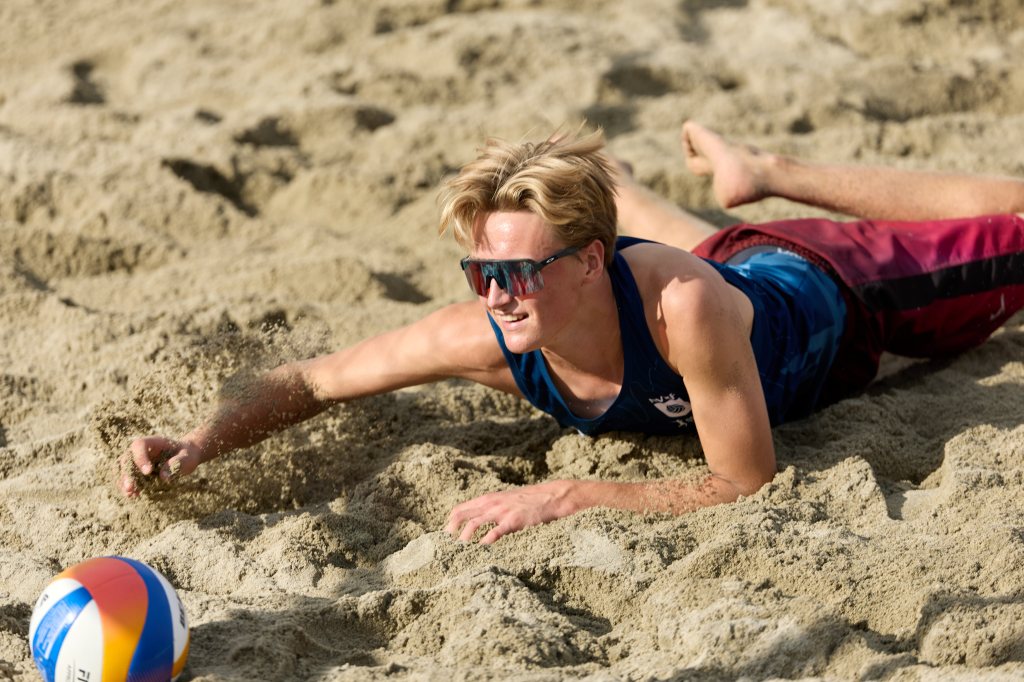 En ung mann i solbriller dykker etter en volleyball på stranden.