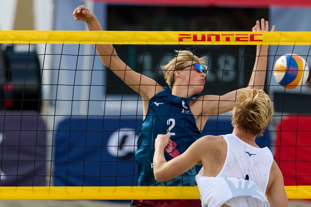 To unge menn spiller volleyball på sand, med en spiller som hopper for å blokkere et angrep mot nettet.